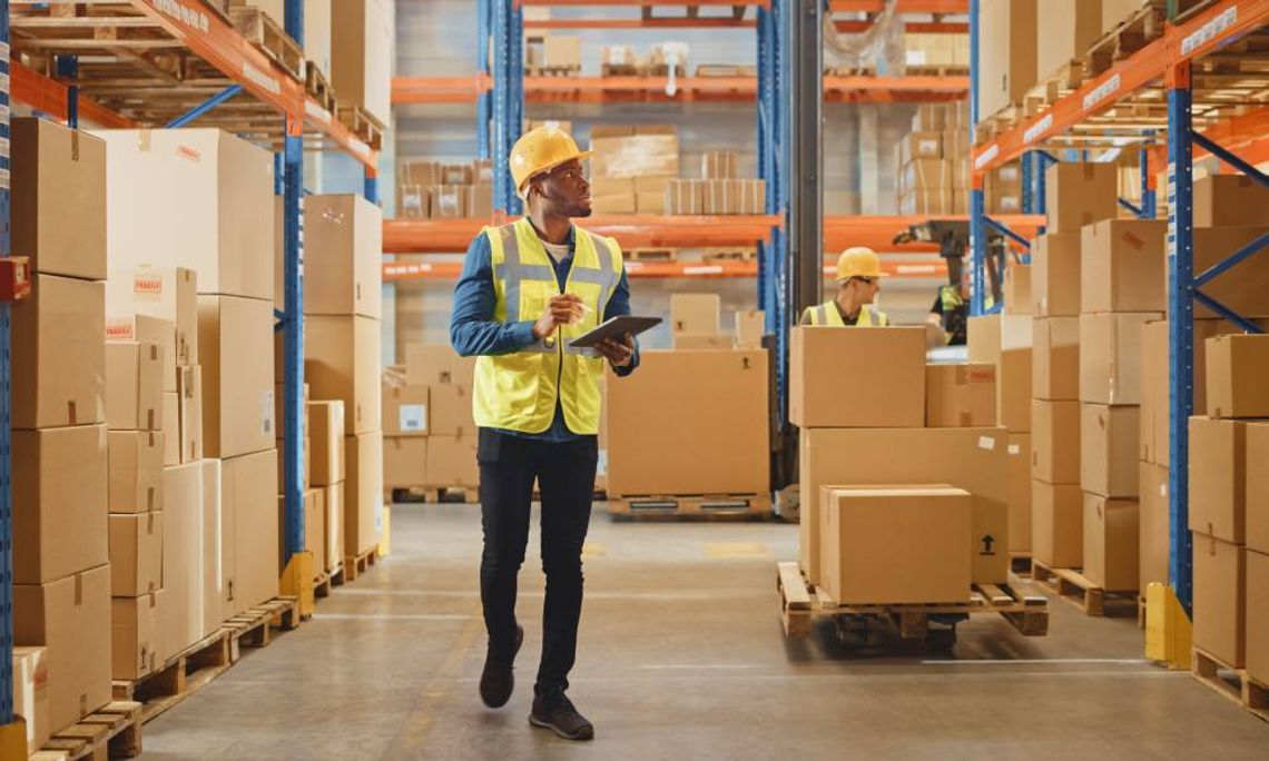 A man in a yellow safety vest and hard hat examining warehouse pallet racking while taking notes on a tablet.