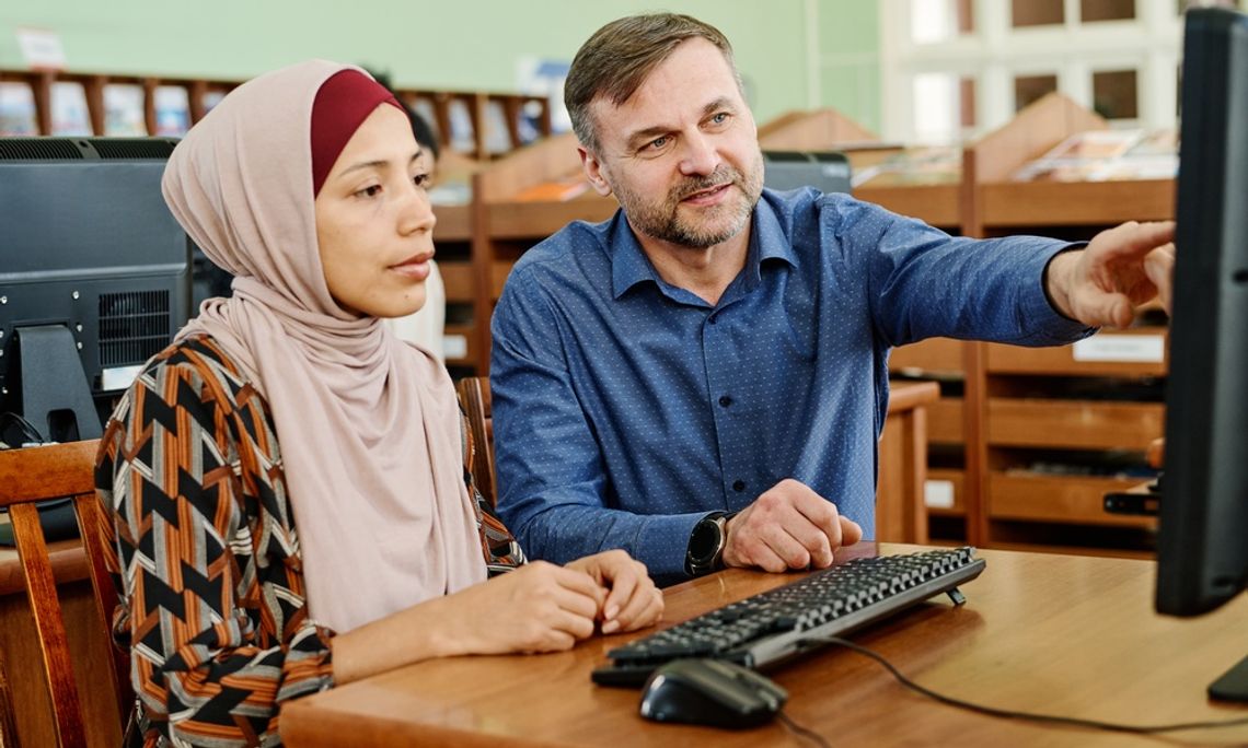 A Caucasian teacher points toward the computer screen while sitting next to a woman wearing a hijab.