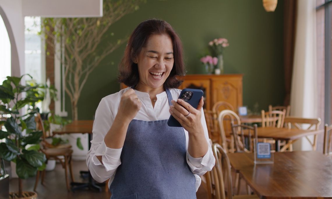 Learning To Master the Art of Restaurant Procurement A smiling restaurant owner in an apron celebrates while looking at her phone inside a cozy, plant-filled café dining area.