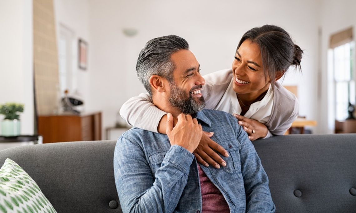 A woman embracing a seated man from behind on a gray couch while they smile at each other in a bright living room.