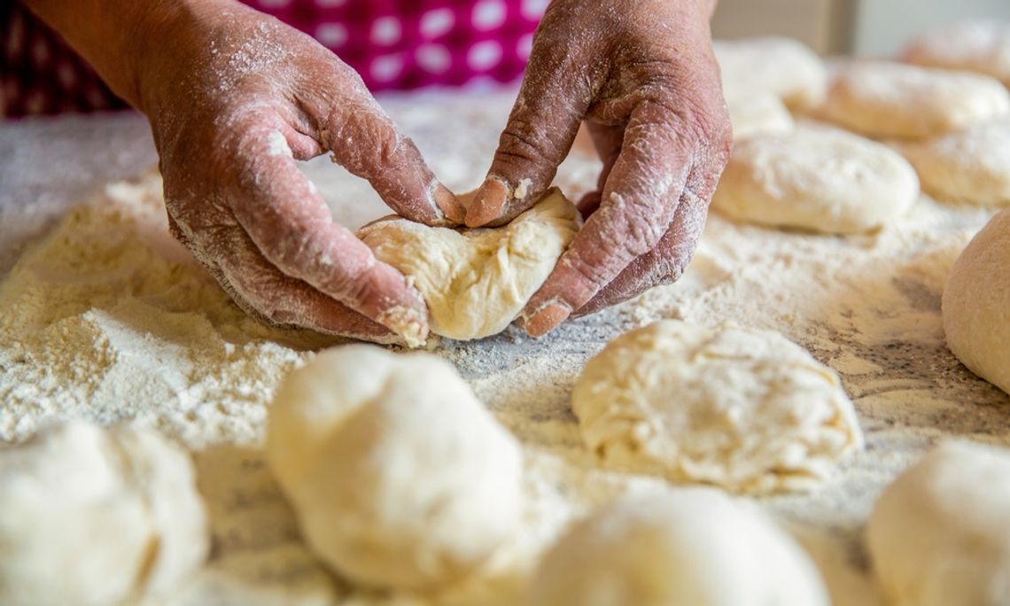 A baker wearing a red checkered apron kneading dough on a floured countertop, surrounded by several other portions of dough.
