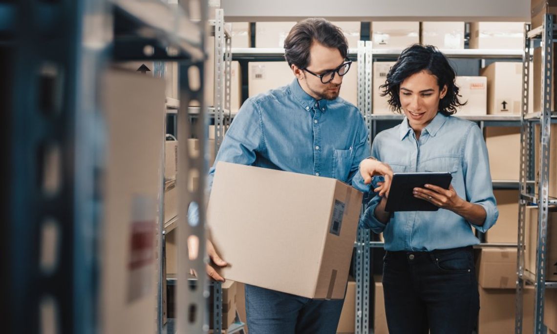 A man and a woman stand in a warehouse looking at a black tablet. The man is holding a cardboard box.