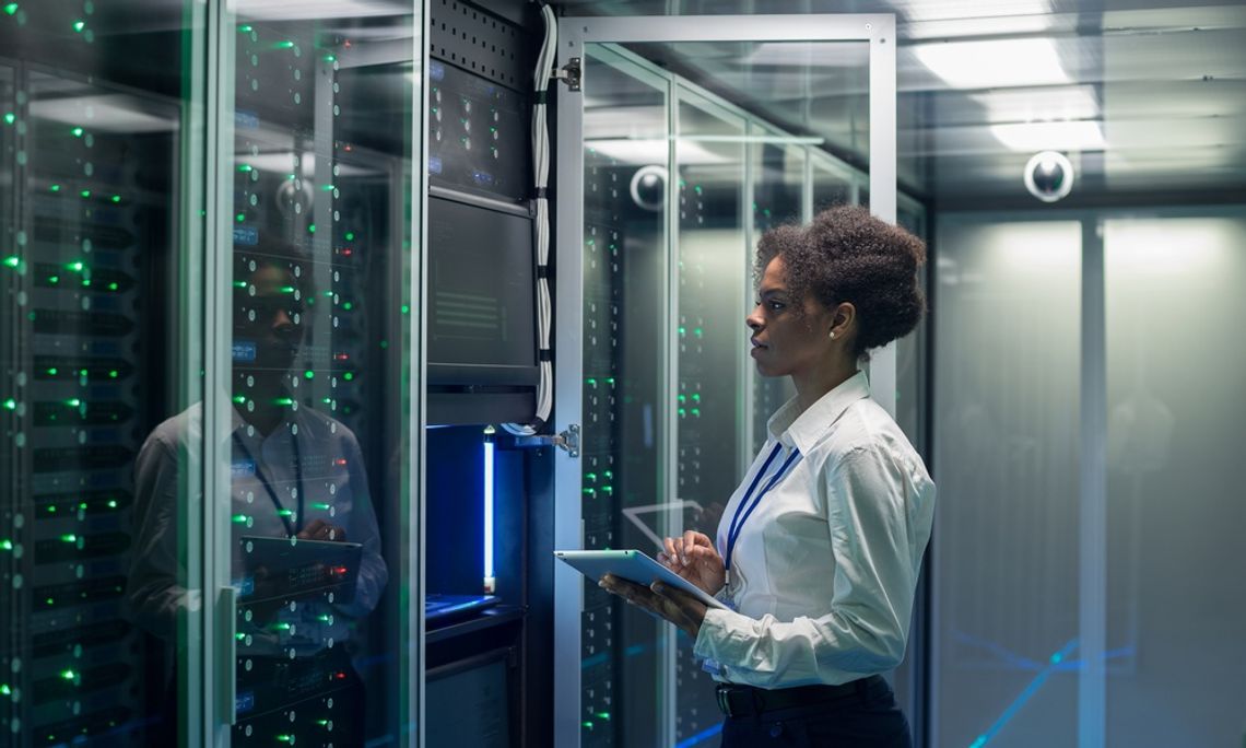A Black data center employee standing in front of an opened server rack with a tablet in her hands.