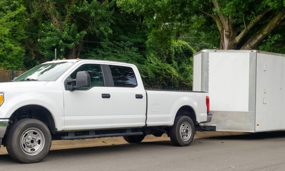 A white trailer attached to a white pickup truck, parked on a street with mature trees in the background.