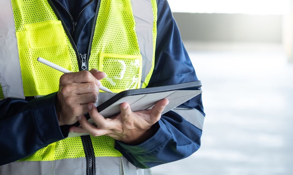 A man in a reflective vest using a digital tablet while inspecting wall-mounted equipment inside a building.