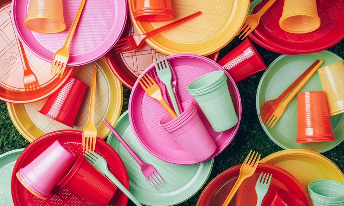 An aerial view shows pink, green, yellow, orange, and red disposable cutlery, plates, and cups scattered across the ground.