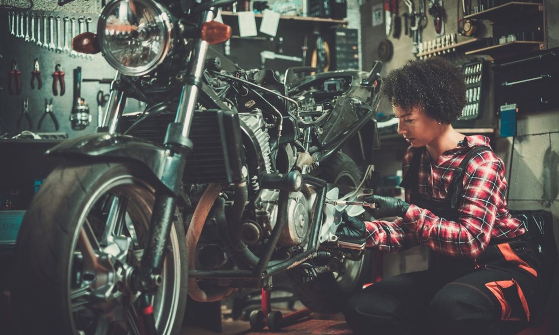 Miles To Go: Preparing for a Long-Distance Motorcycle Ride A young woman wearing bib overalls kneels next to a motorcycle and turns a torque wrench. Tools hang on the walls.