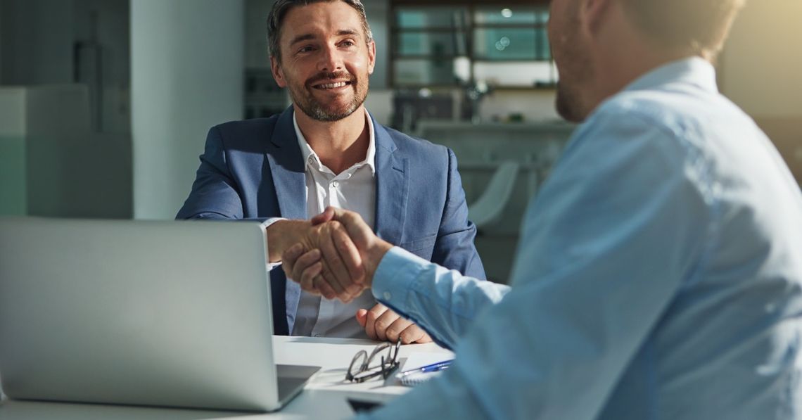 Mistakes Interviewers Make When Talking to Candidates Two professionally dressed employees shaking hands over a large table with a laptop and glasses on it.