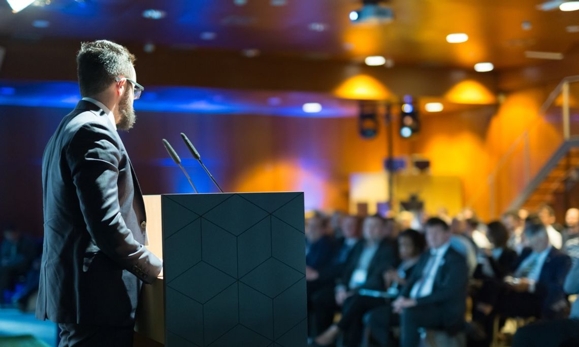 A guest speaker standing on the stage at a corporate event. The crowd is full of professionally dressed attendees.