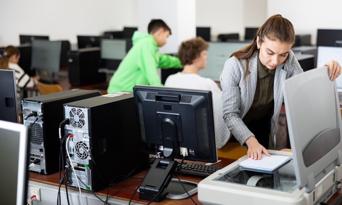 Printer Maintenance Tips for Teachers and Schools A woman in a gray blazer placing papers on a flatbed scanner in a computer classroom with students seated behind her.