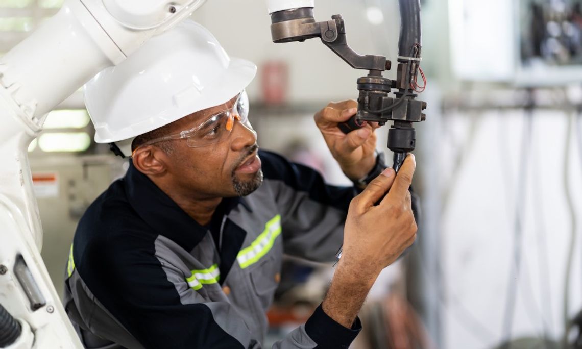 Protecting Investments: Best Practices for Machinery Care A man wearing a hard hat and safety glasses adjusts two levers on a robotic arm in a brightly lit setting.