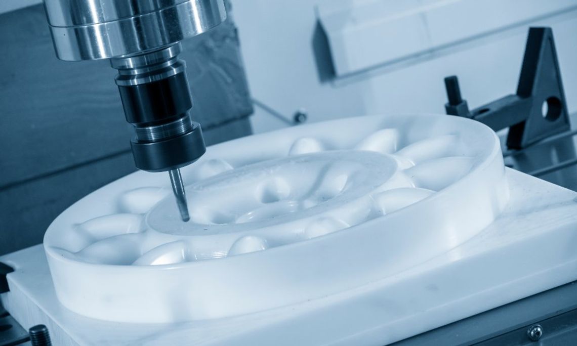 Close-up of a milling bit cutting a smooth white round part on a CNC machine bed in a workshop with cool blue lighting.