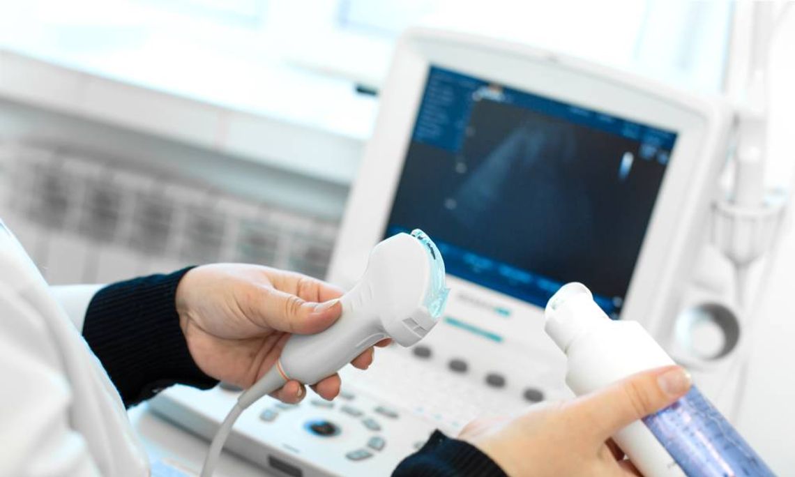 A medical professional applies a blue gel to the end of an ultrasound transducer. The ultrasound machine appears blurry.