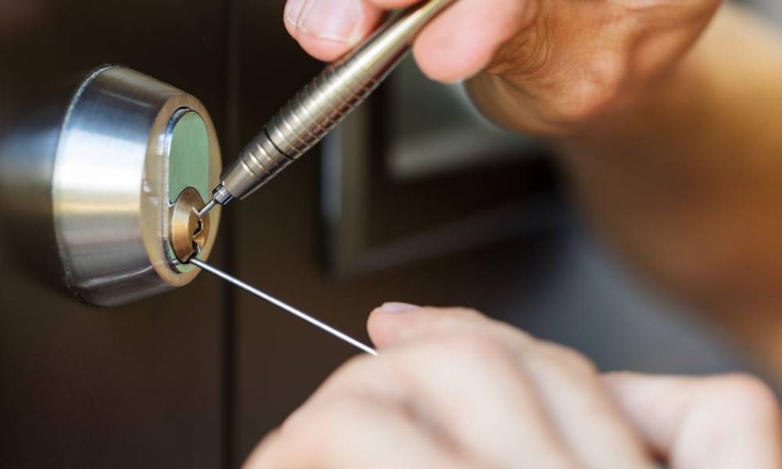 A close-up view shows two hands placing silver lock picking tools into a silver lock on a brown door.