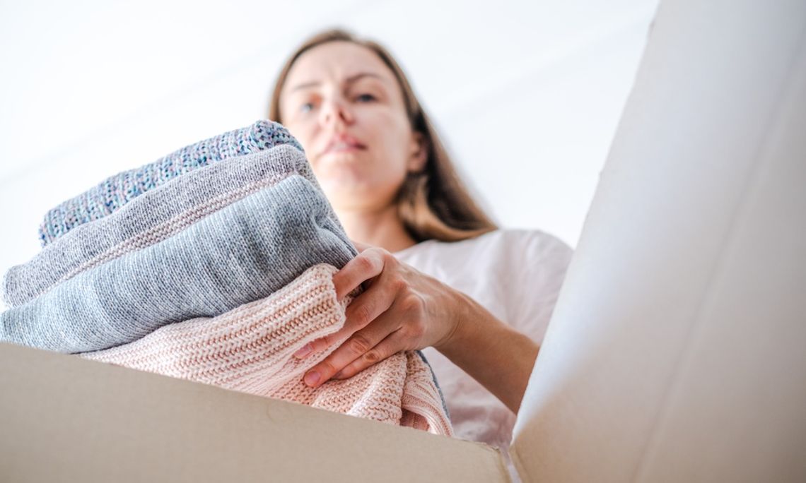 Reasons To Rent a Roll-Off Dumpster for a Home Cleanout A low-angle view of a woman placing a pile of folded blue and pink sweaters into a brown cardboard box.