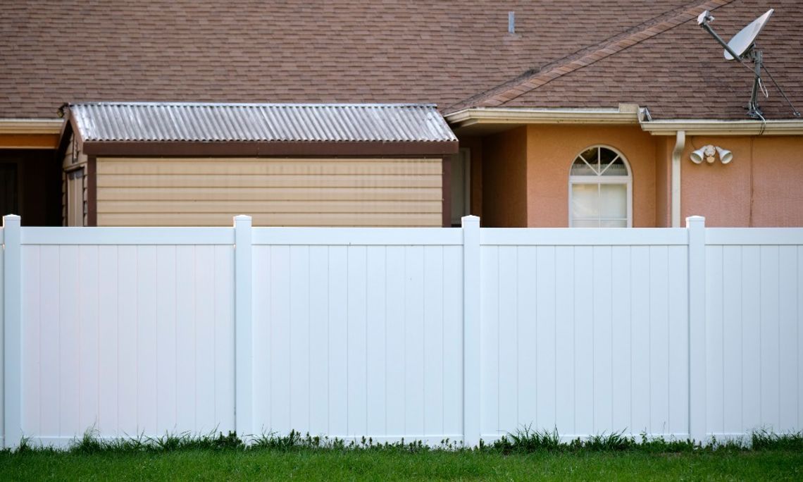 A white PVC fence installed in front of a peach-colored home with a metal shed in its yard during the daytime.