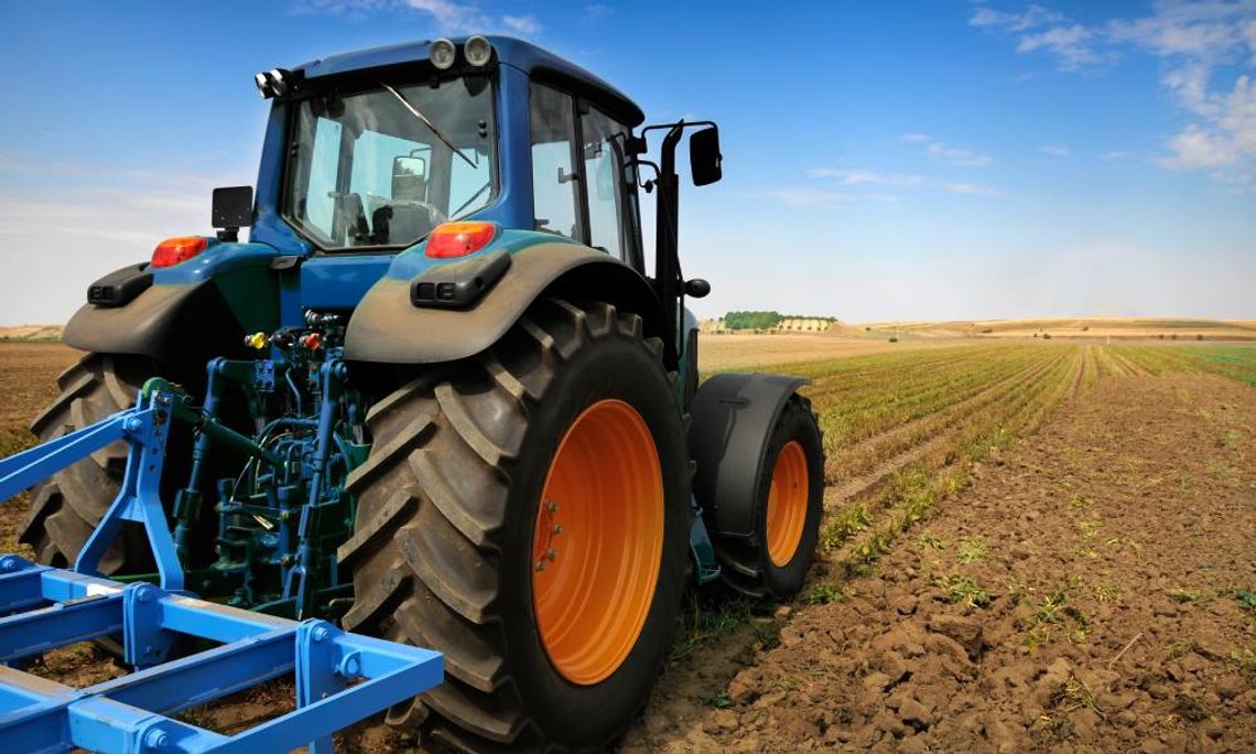 Reducing Downtime in Industrial and Farm Equipment Blue tractor working on a cultivated field with rows of crops under a clear blue sky in a rural farming landscape.