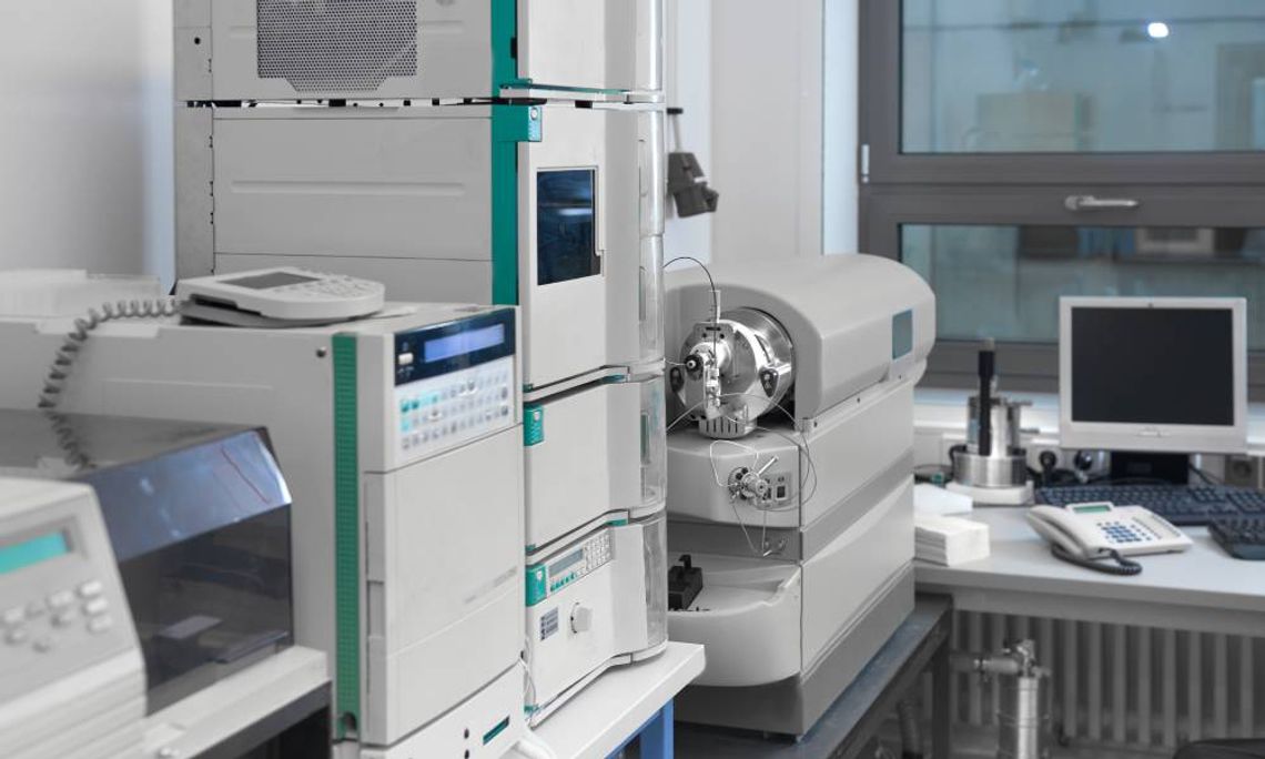 A laboratory interior with several white and teal machines on a countertop near a desk with a monitor and keyboard.