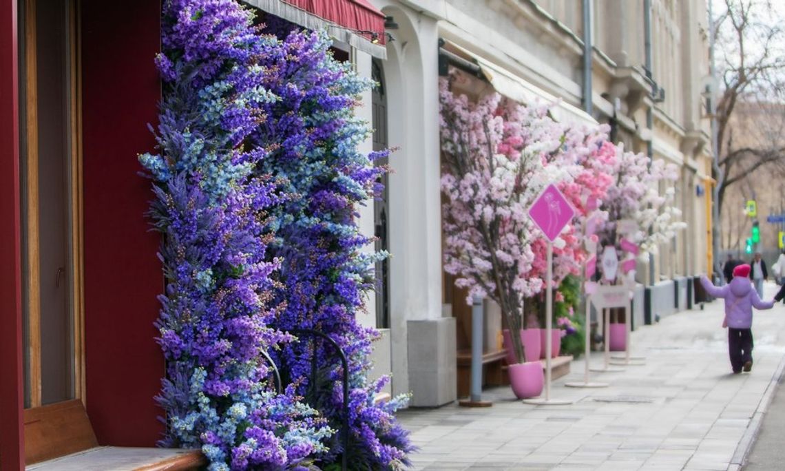 People walk past storefronts. Purple and blue flowers surround one doorway, and pink and white flowers surround the next.