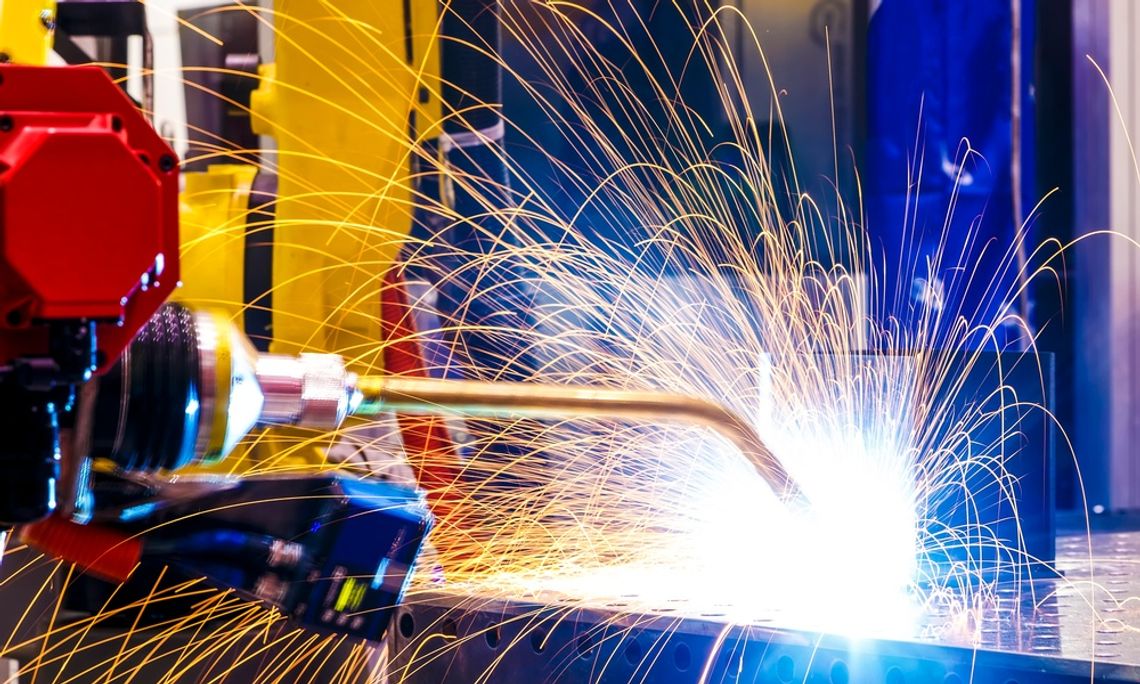 A yellow robotic arm welds a rectangular metal part, emitting yellow-orange sparks from the point of contact.