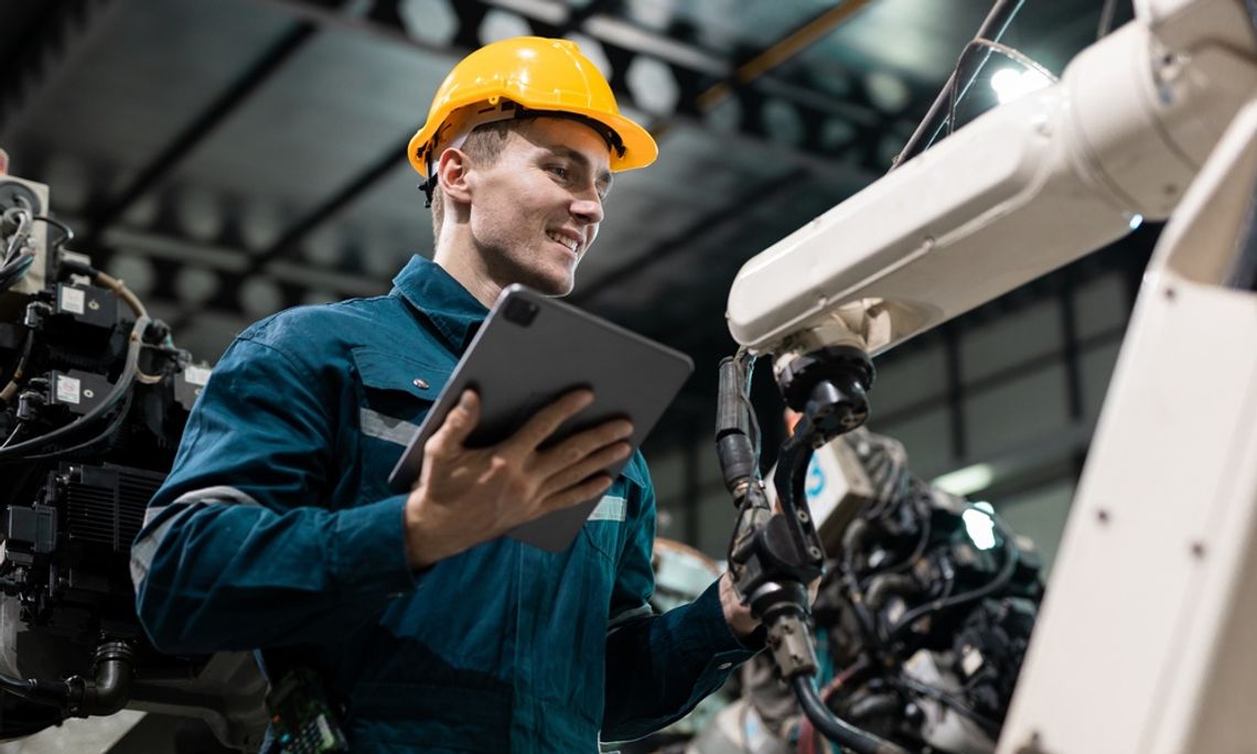 A factory worker with a tablet and hard hat is examining a robotic arm on an automated production line system.