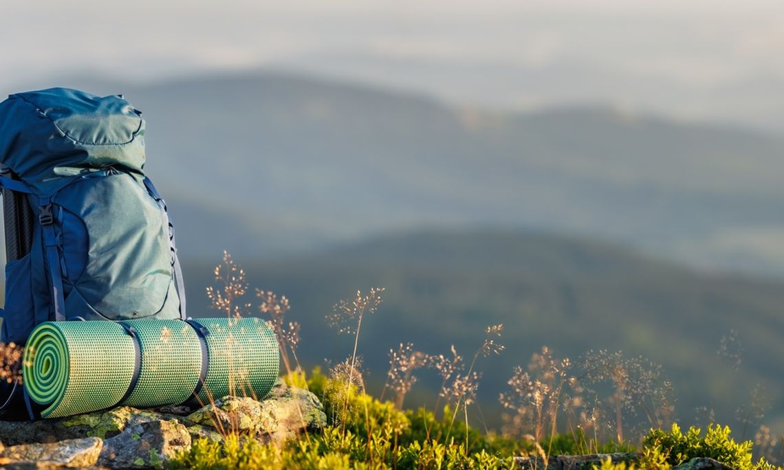 A blue hiking backpack and rolled sleeping pad resting on rocks and grass with blurred mountains in the background.
