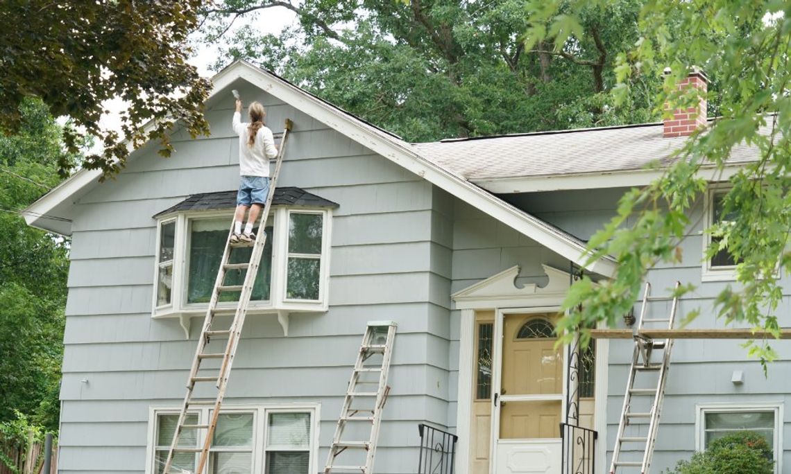 Person on a ladder paints the upper siding of a gray house, with extra ladders and trees around the yard.