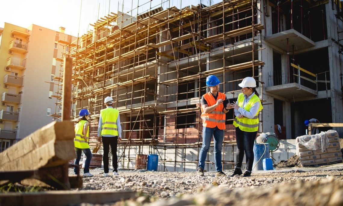 Construction workers stand around an active build site with safety vests and hard hats on. Two are talking about plans.