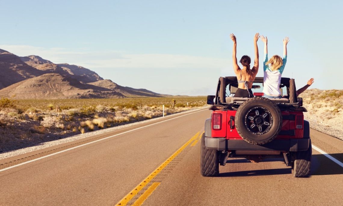 A red vehicle driving on a desert road with passengers raising their arms near mountains under a clear sky.