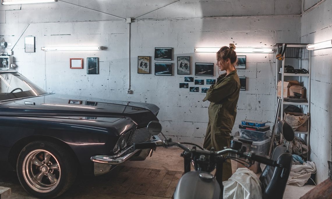 Spark Plug Basics for Weekend Car Projects A blonde woman in a mechanic suit stands with her arms crossed in front of a vintage car parked in a garage.