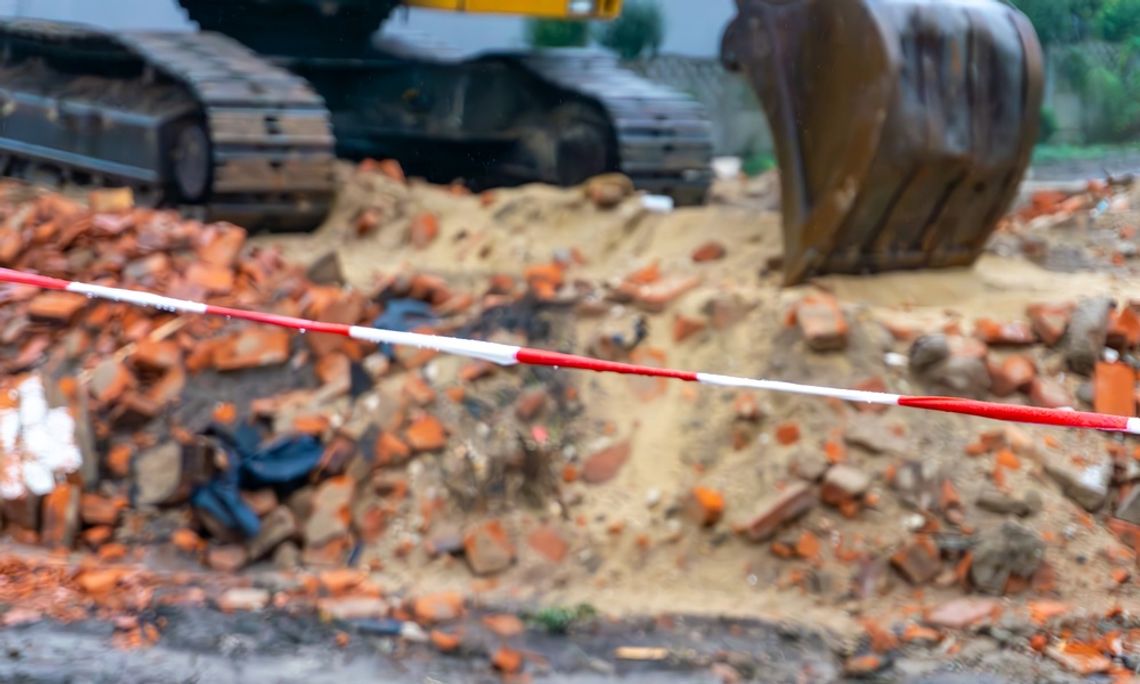 A crawler excavator is poised above a mound of dirt and bricks behind a line of red-and-white warning tape.