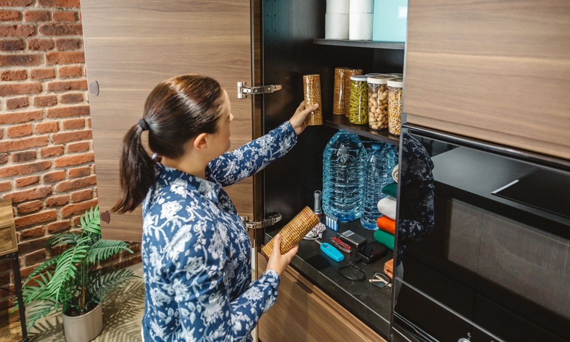 A woman organizes a modern kitchen pantry with jars, bottled water, and emergency tools, set against a brick wall.