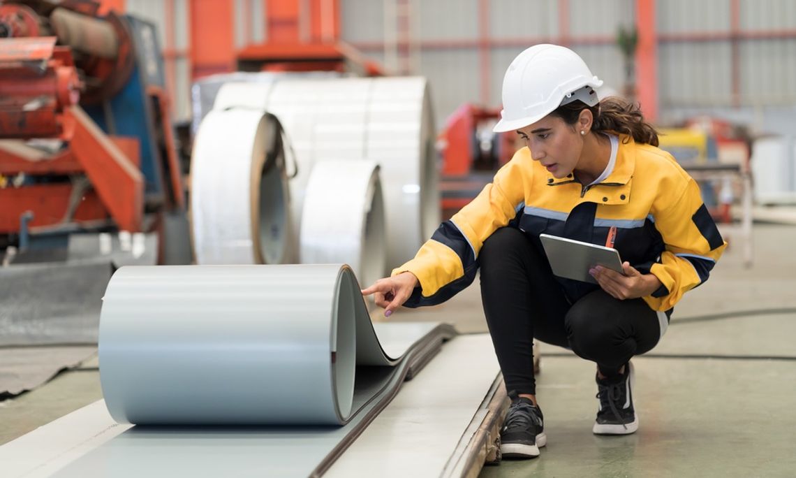 A factory worker is inspecting the quality of a metal sheet. She is in a factory wearing safety gear.