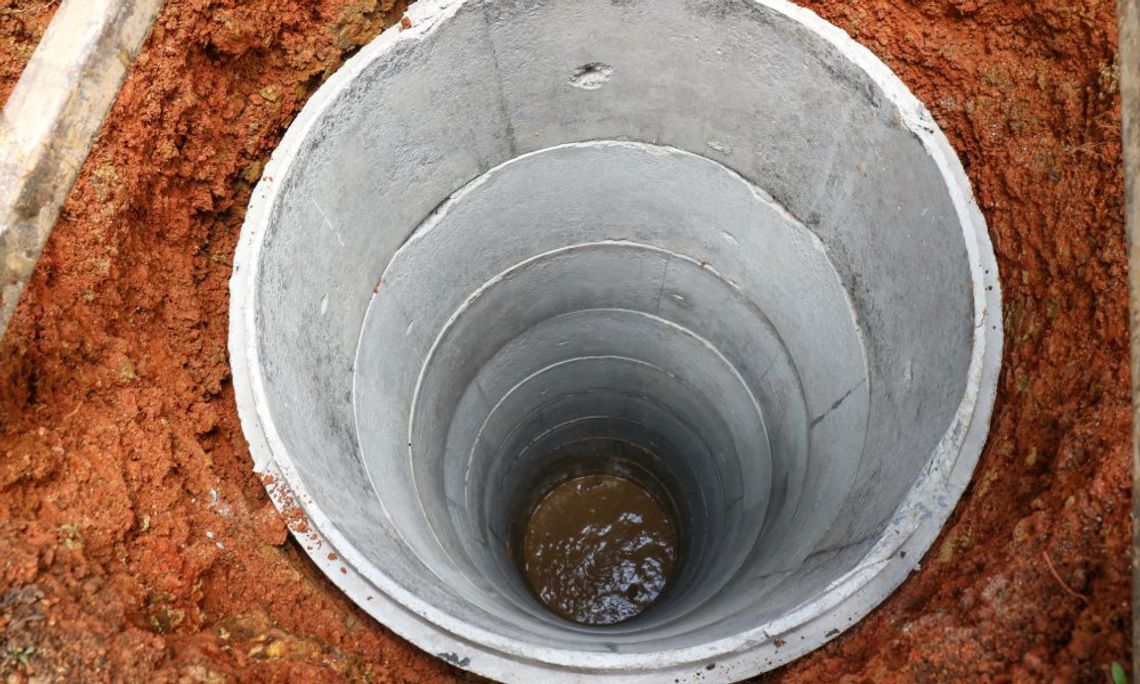 A bird's eye view shows a concrete well filling up with water and surrounded by orange clay mud.