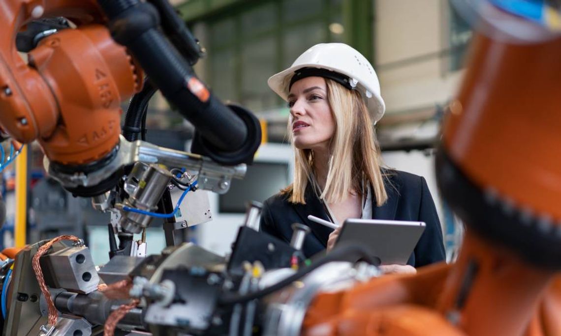 A professional woman wearing a white hard hat holds a tablet while she examines the components of a machine.