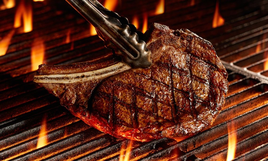 The Best Steak Cuts for Southern-Style Grilling Tongs lift a Bone-In Ribeye steak as it grills over open flames, showing perfect char marks and a caramelized crust.