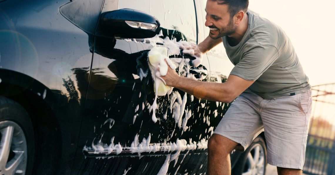 The Best Ways To Keep Your Vehicle Looking Good for Years A close-up of a man smiling while using a sponge with soapy water to clean the exterior of a black vehicle.