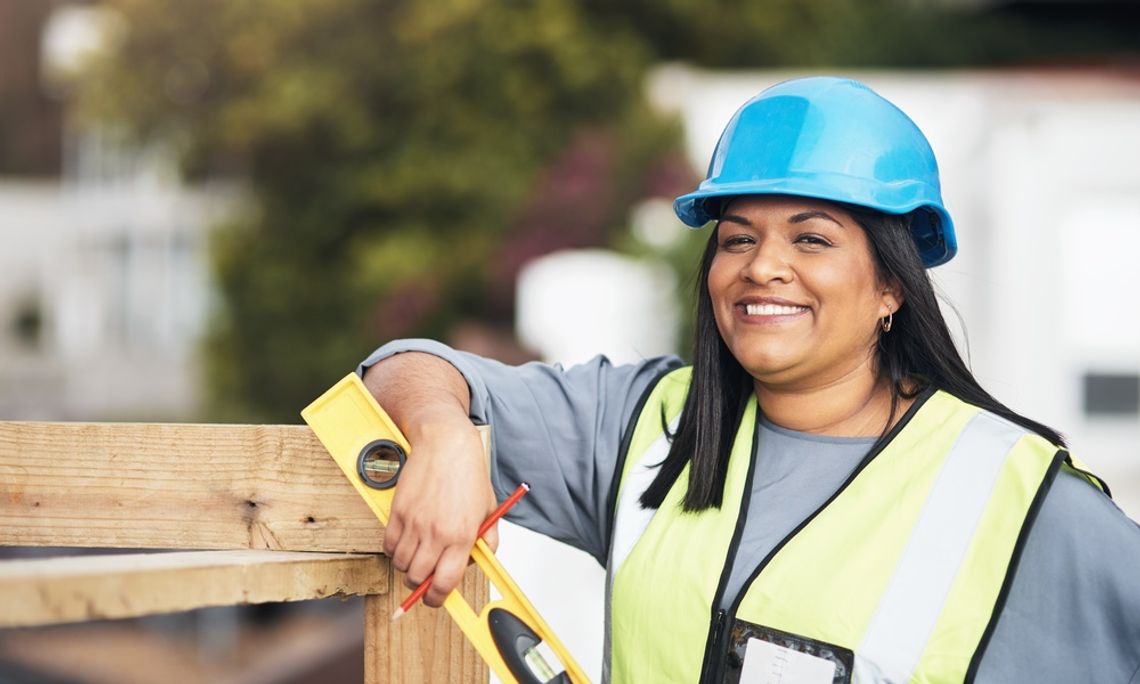 A construction worker wearing a safety vest and a hard hat leaning on a wooden structure with a level in her hand.