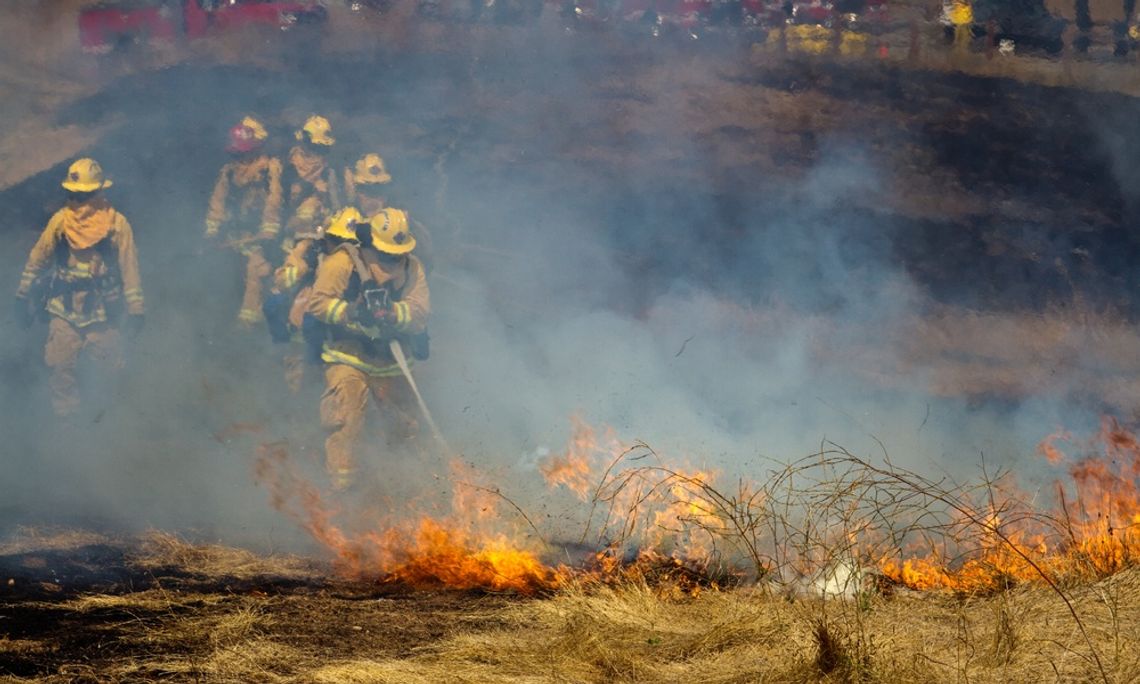 A group of five wildland firefighters wearing high-visibility protective gear stand in a line while holding a hose.