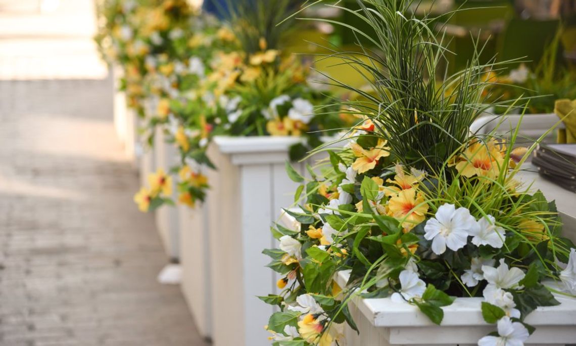 A series of white pillars with yellow and white flowers in front of a restaurant patio and stone street.