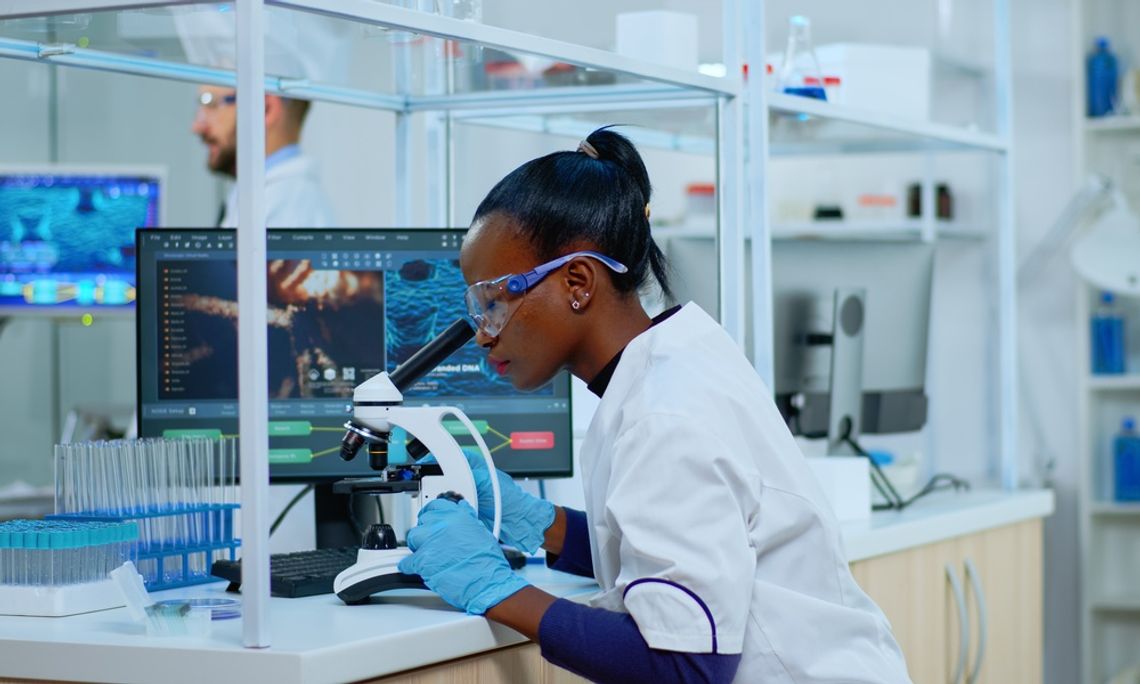 A young Black female scientist checking sample of virus using microscope in modern lab. She's wearing safety gear.