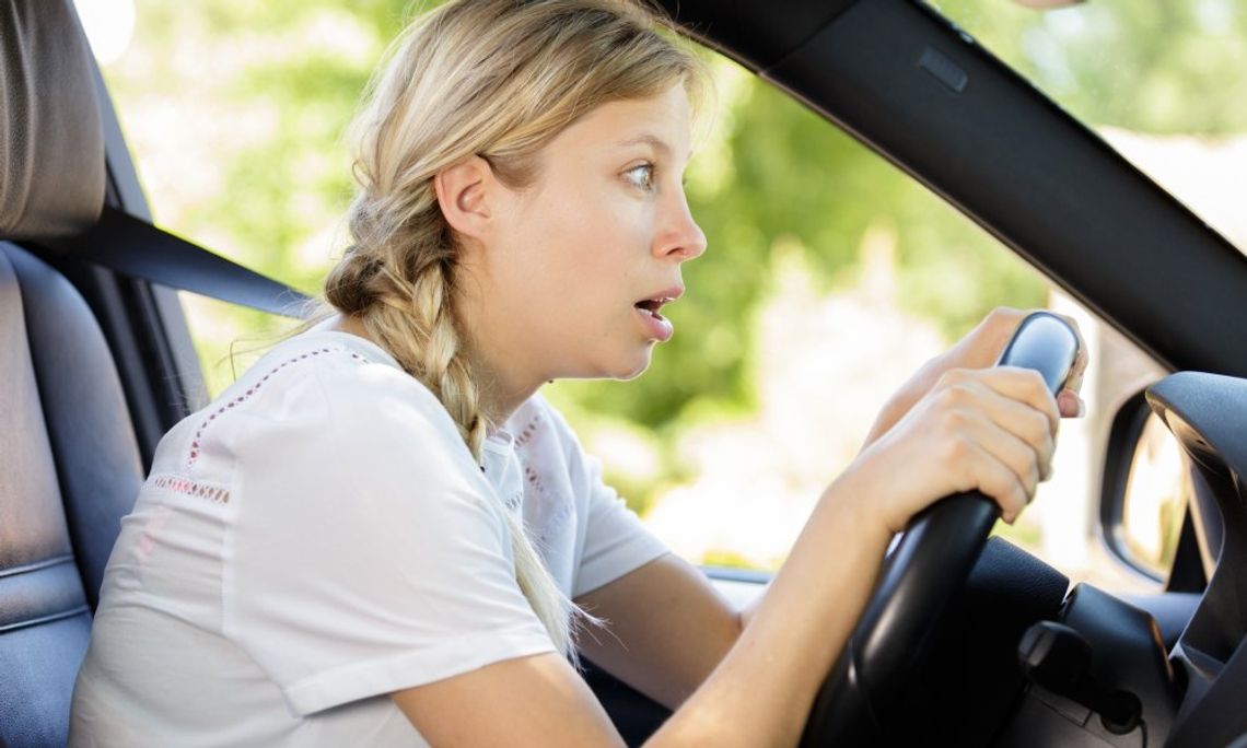 A blonde woman looks shocked as she grips her car's steering wheel and stares wide-eyed through the windshield.