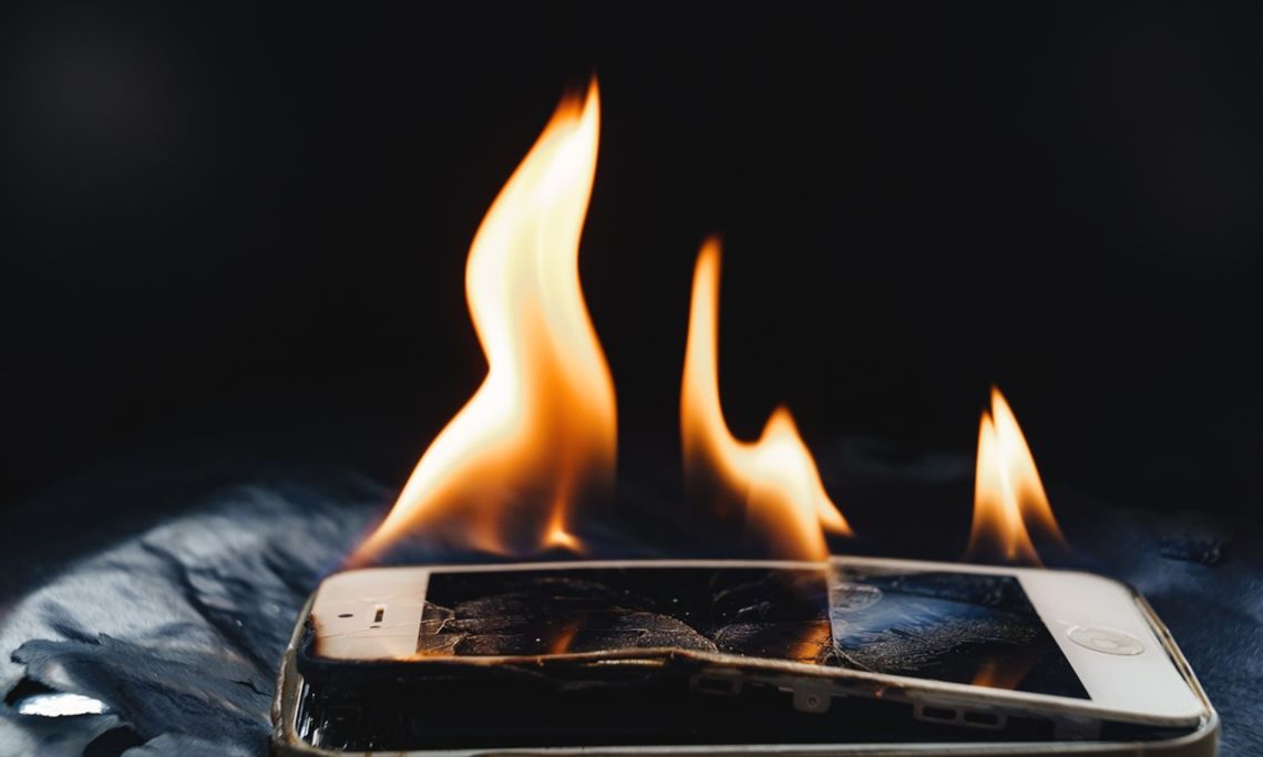 A cell phone on fire with a cracked, bent screen with smaller flames. The screen is splitting on the edge.