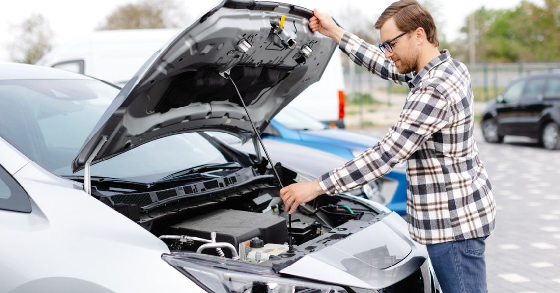 A person wearing glasses opening the hood of a gray car parked outside next to a blue car during the daytime.