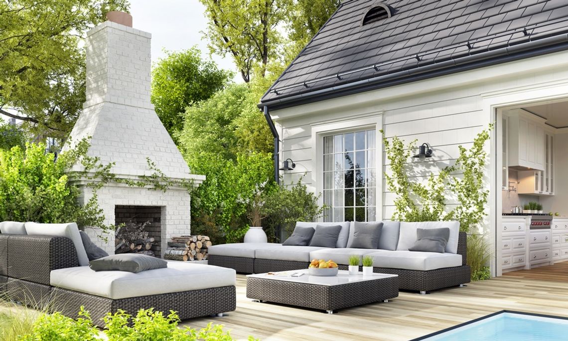 The Most Useful Luxury Features To Add to Your Home Luxury backyard patio with modern outdoor seating, a white brick fireplace, lush landscaping, and an open view into a stylish kitchen.