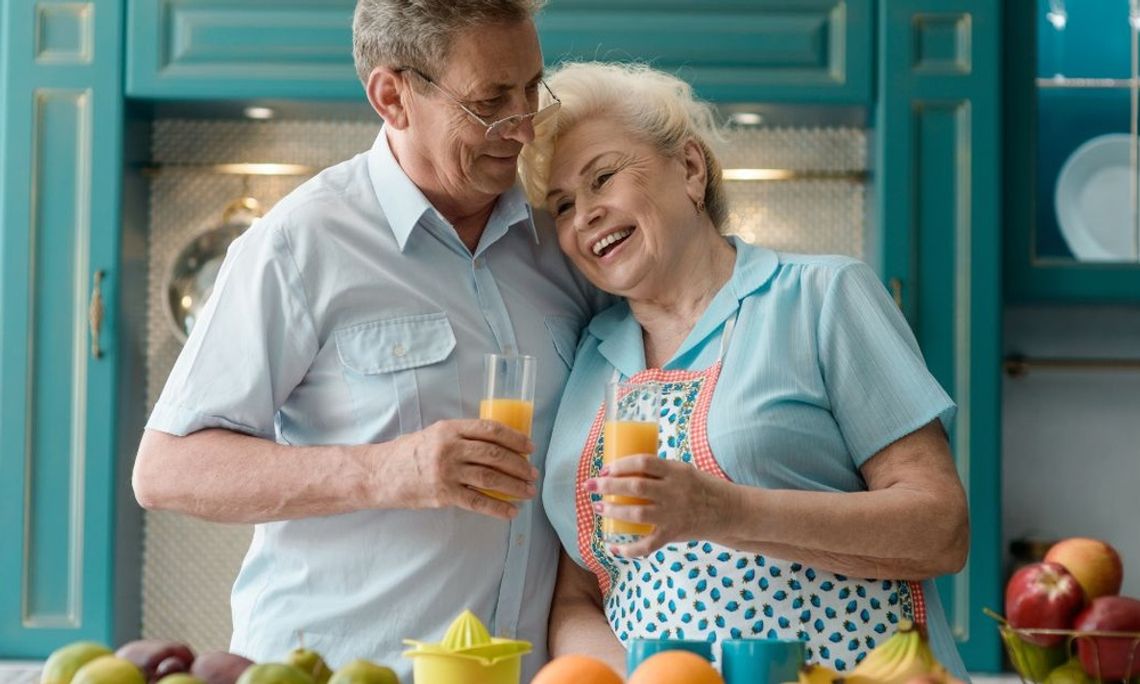A senior couple is leaning against each other while holding cups of juice. A counter with produce is in front of them.