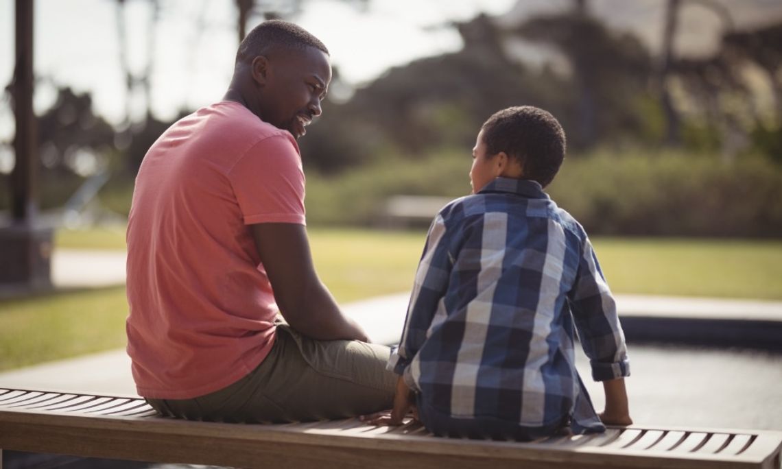A man sits on a wooden bench next to an elementary school-aged boy at a park. They are facing each other.