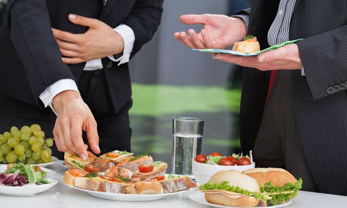 Two business professionals wearing nice suits stand at a lunch buffet in their office. One reaches to pick up a small sandwich.
