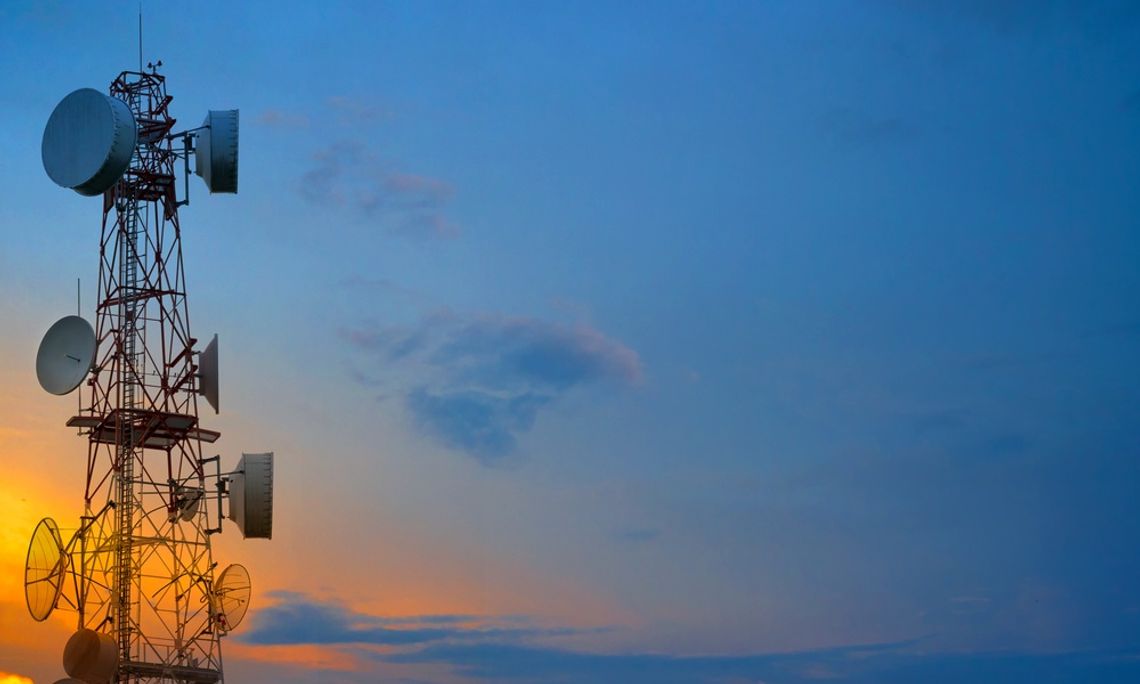 Two telecommunication towers with wireless antennas against with the sun setting behind clouds behind them.