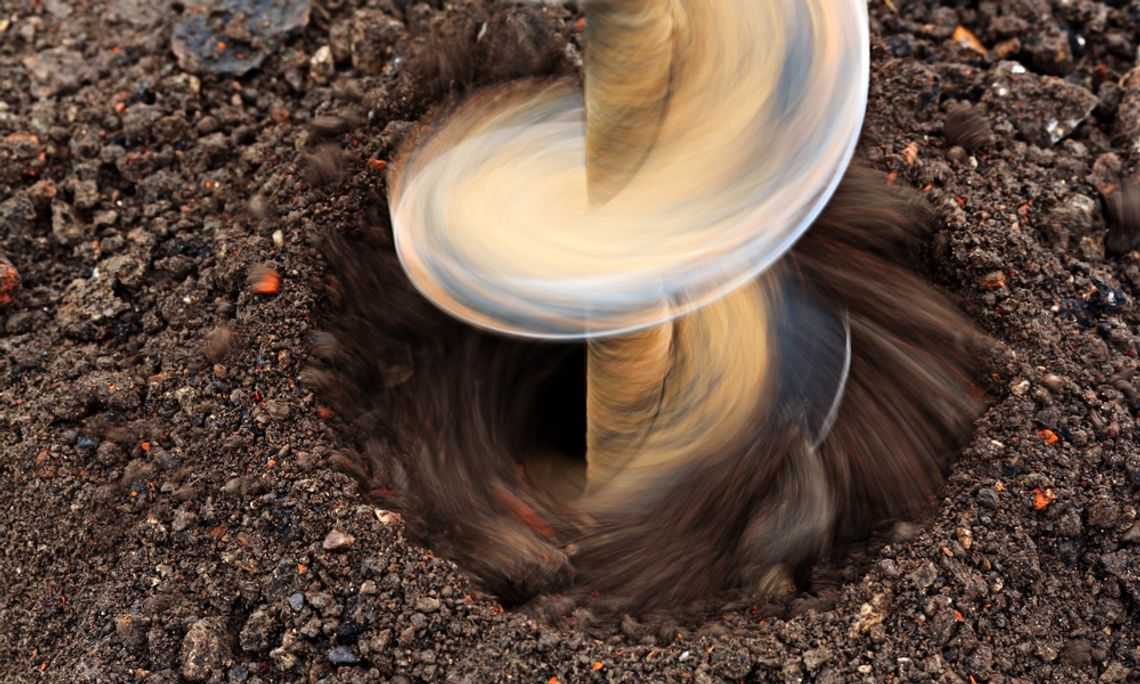 Close-up of a rotating earth auger drilling into dark soil, creating a deep hole as loose dirt spins around the bit.